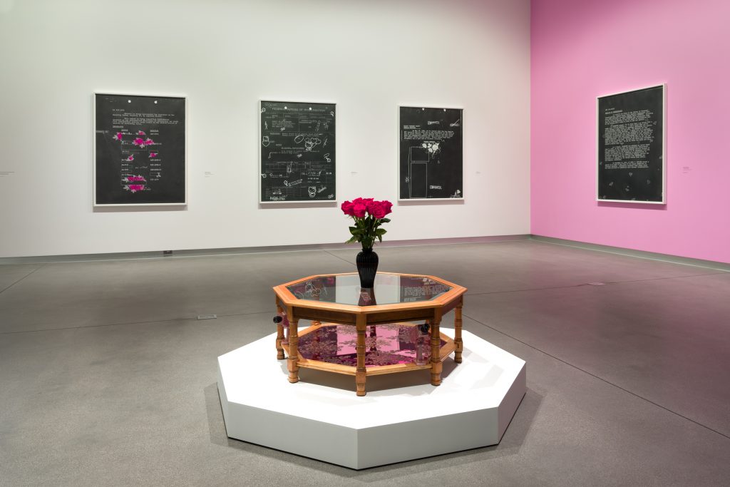 Gallery with one white wall and one pink wall. In the center is a wooden table with a vase of roses