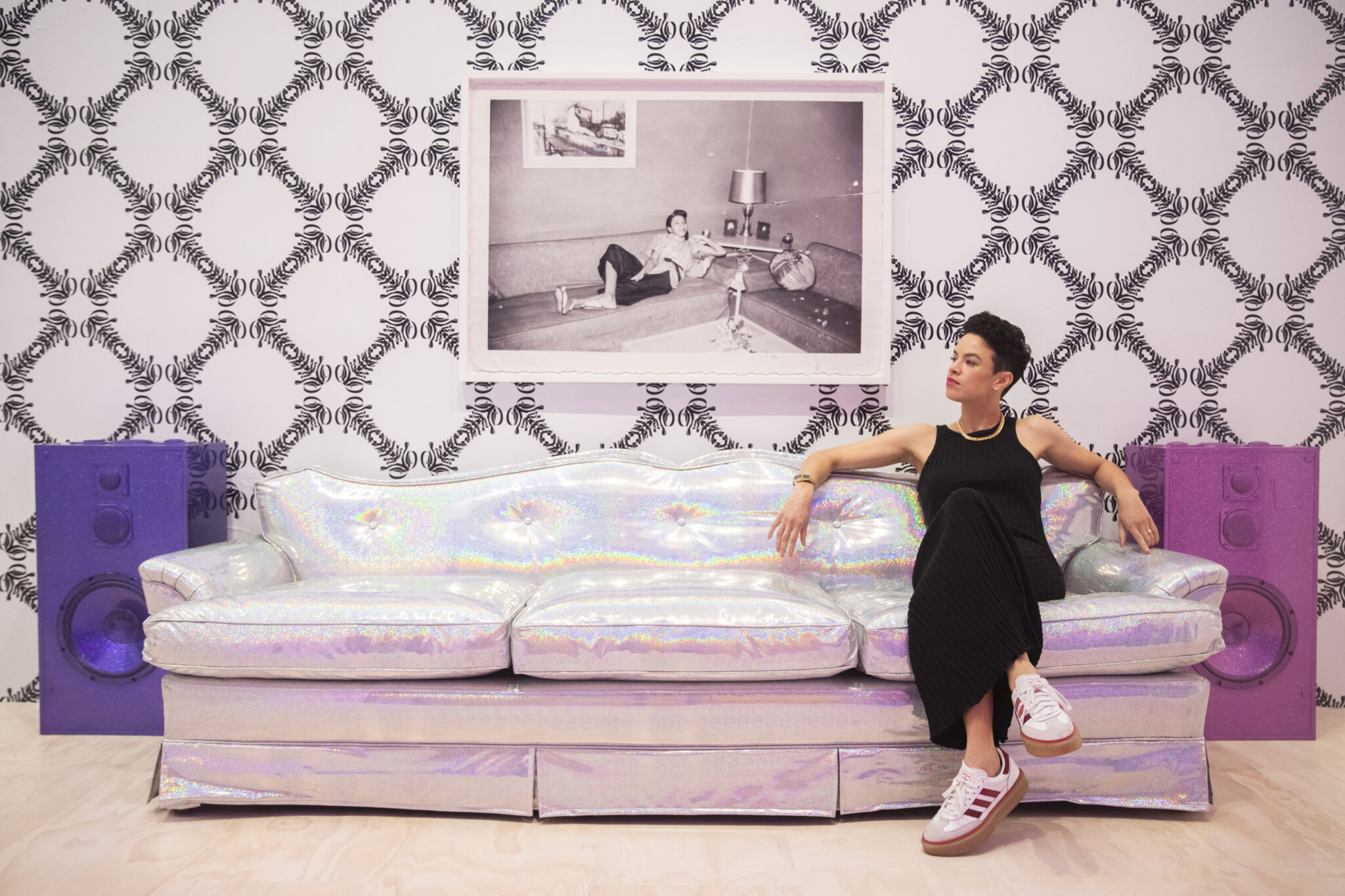 Sadie Barnette sits on her silver holographic couch in a wallpapered room. A large photo is framed behind her. A purple and a pink glittery speakers are on either side of the couch.