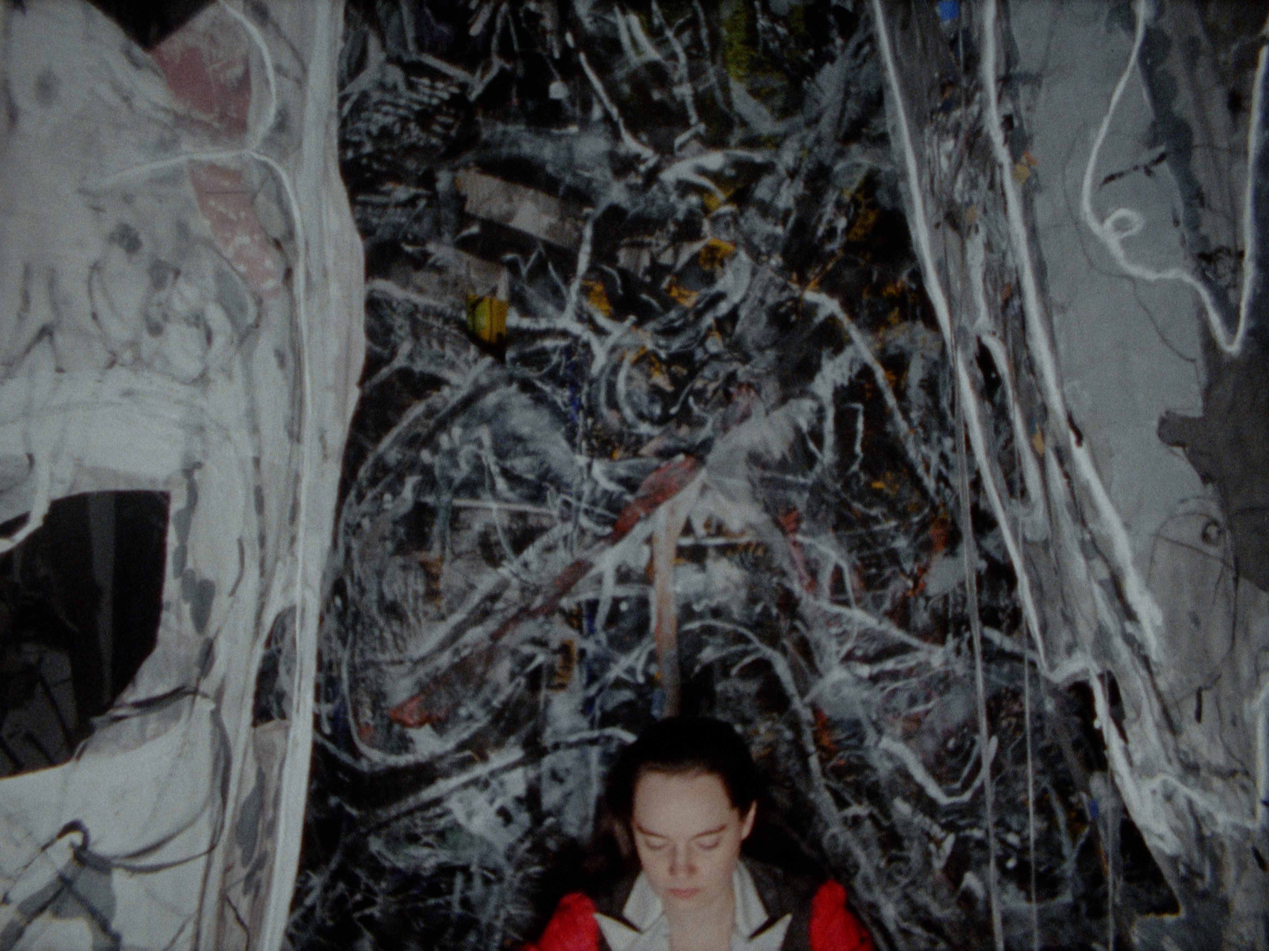 woman in red jacket sits in front of abstractly painted dark canvases