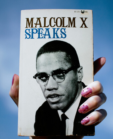 Hand holding up book of 'Malcolm X Speaks' with pink nailpolish against blue backdrop