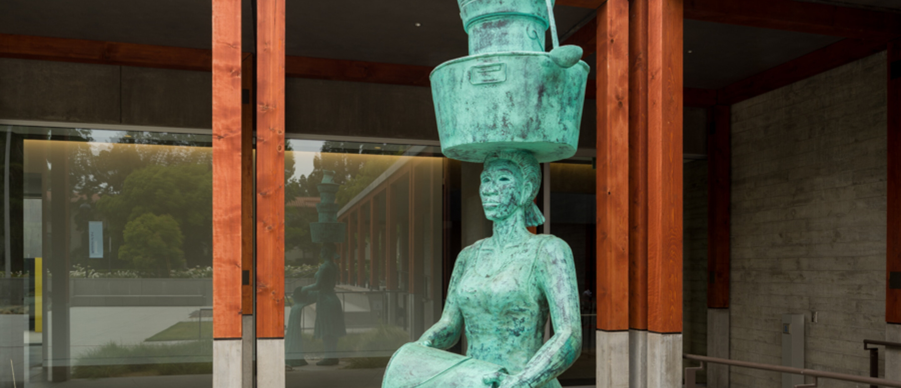 Tall bronze outdoor sculpture of female figure holding stacked pots above while pouring water from a handheld pot