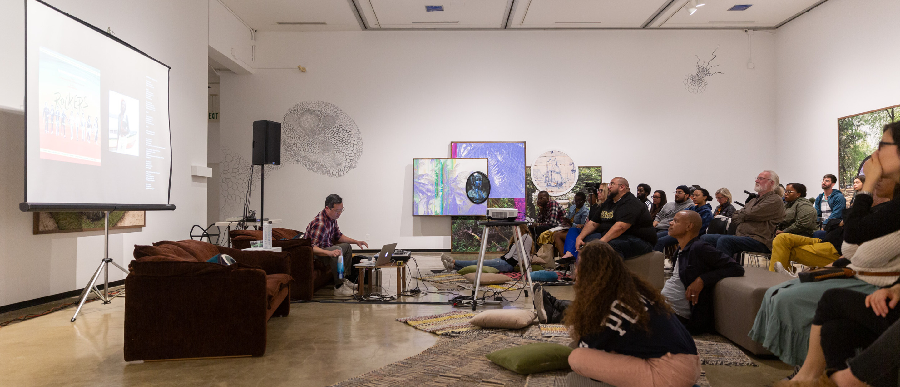 Wideview of an event in a gallery with speaker up front and group of people in the audience