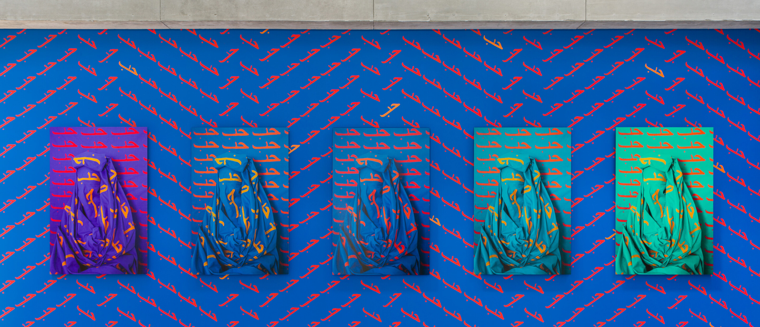 Installation view of Alia Ali's site-specific installation of Love with blue painting wall with Love written repeatedly in Arabic with 5 photographs of figures draped in fabric also with the word Love written in Arabic repeatedly