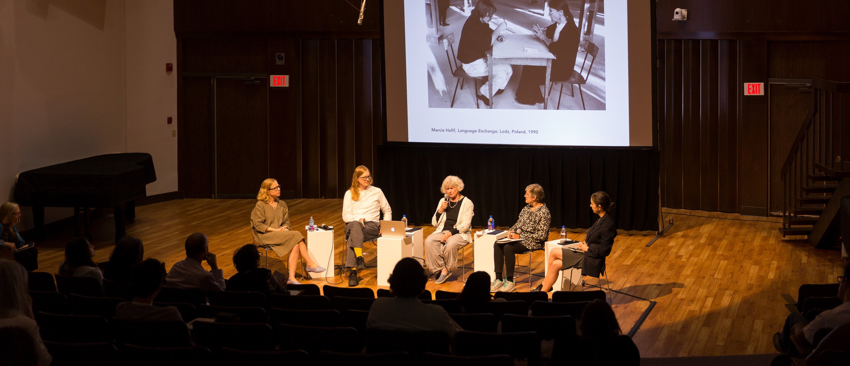 Panel of 5 people in front of a projection screen with an auditorium filled with audience members