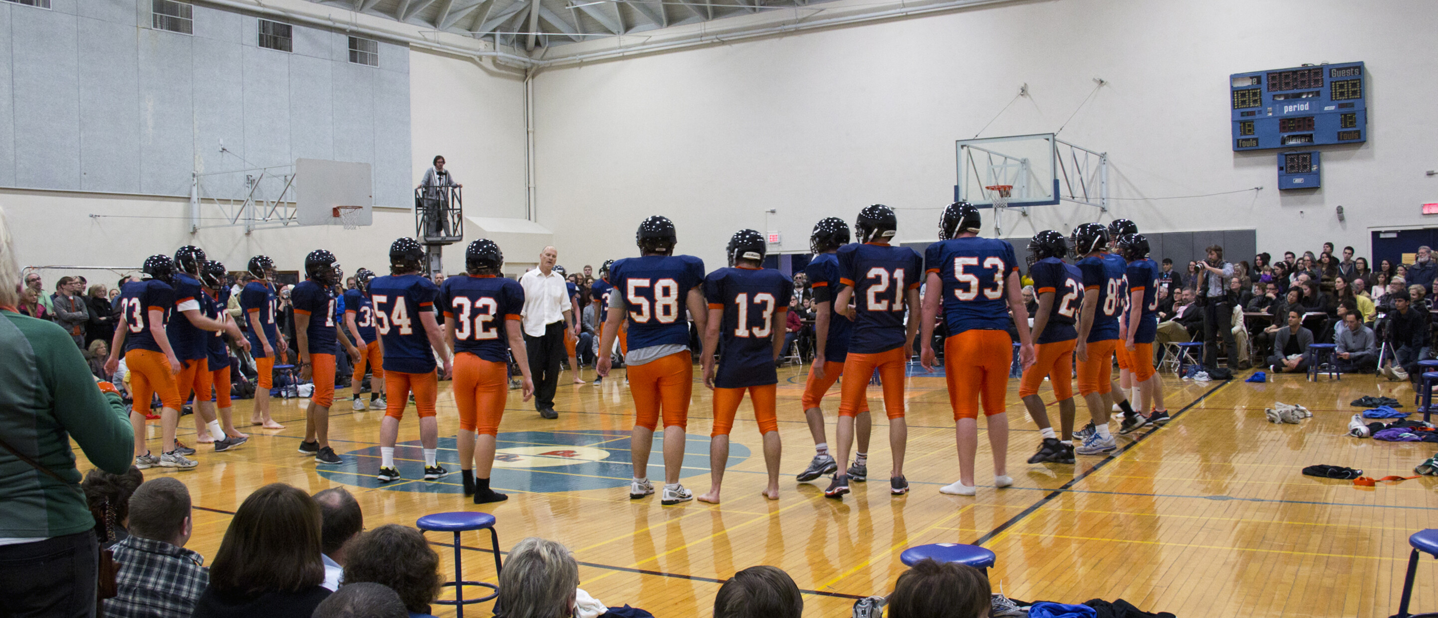 Inside of a gym with a large group of audiences with a football team in the midst of a performance 