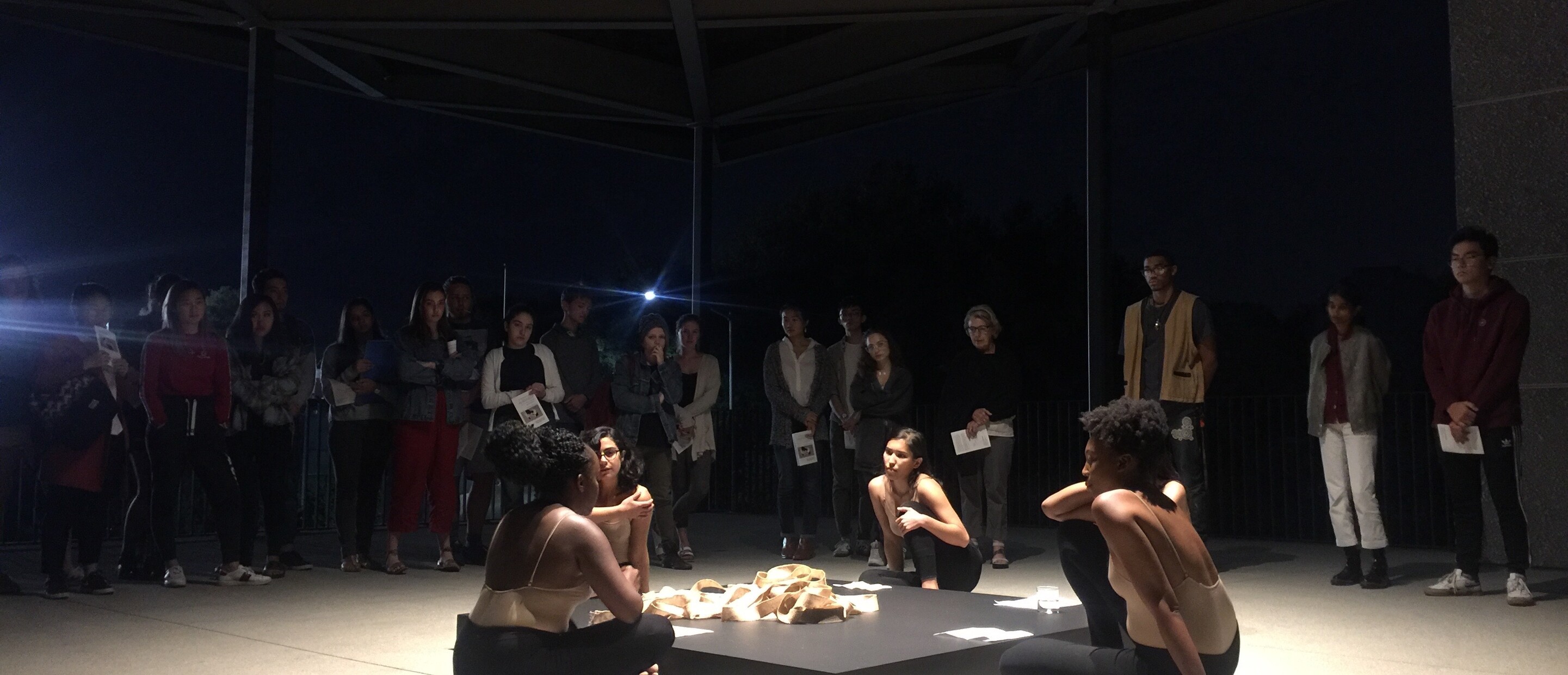 4 women sitting around a square block with an audience behind them during a performance