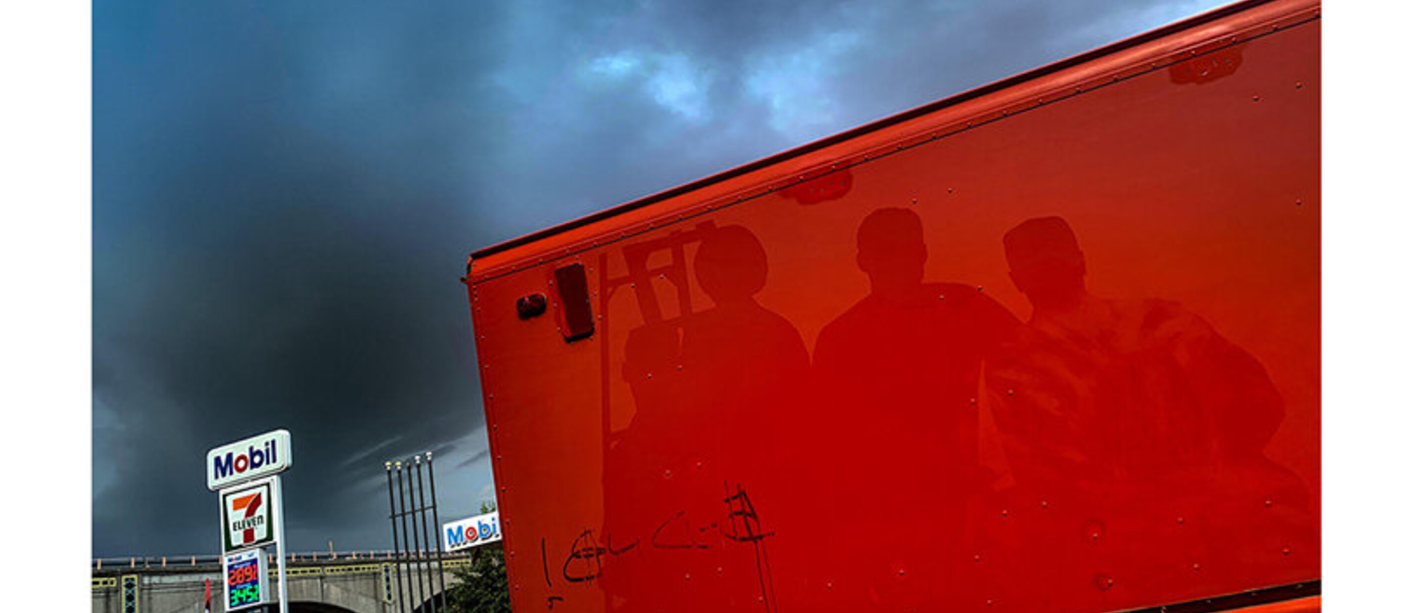 Reflections of individuals against red surface outdoors near gas station
