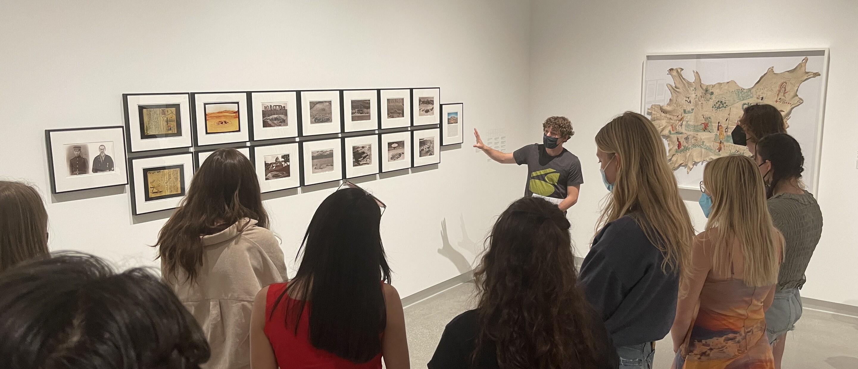 student gestures towards photographs on gallery wall in front of another group of students