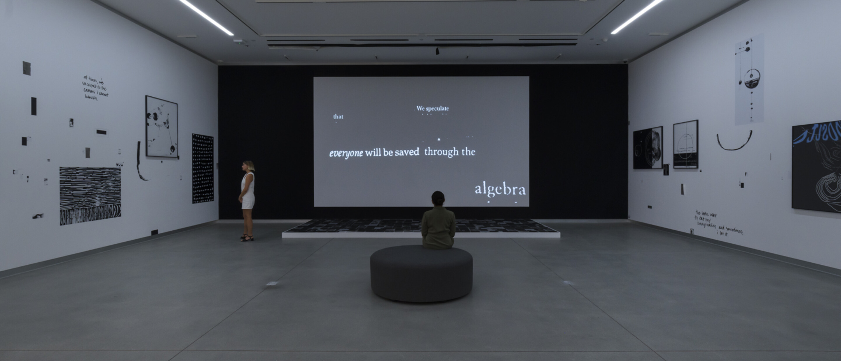 dark room with black and white works and video. two people sand and sit in the room