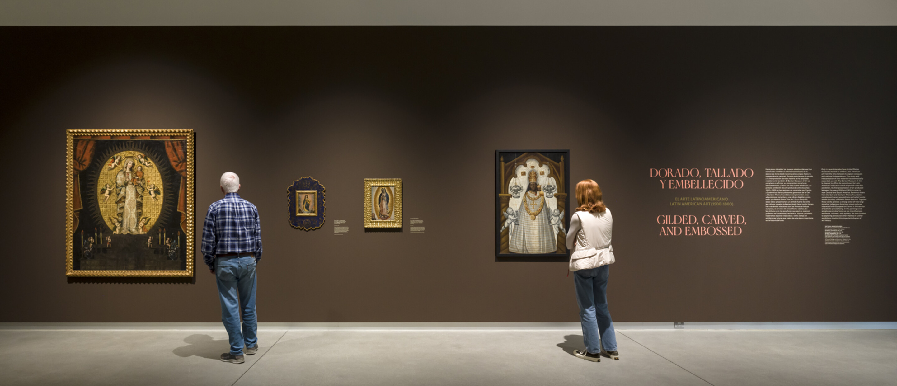 two people look at brown gallery wall with multiple paintings