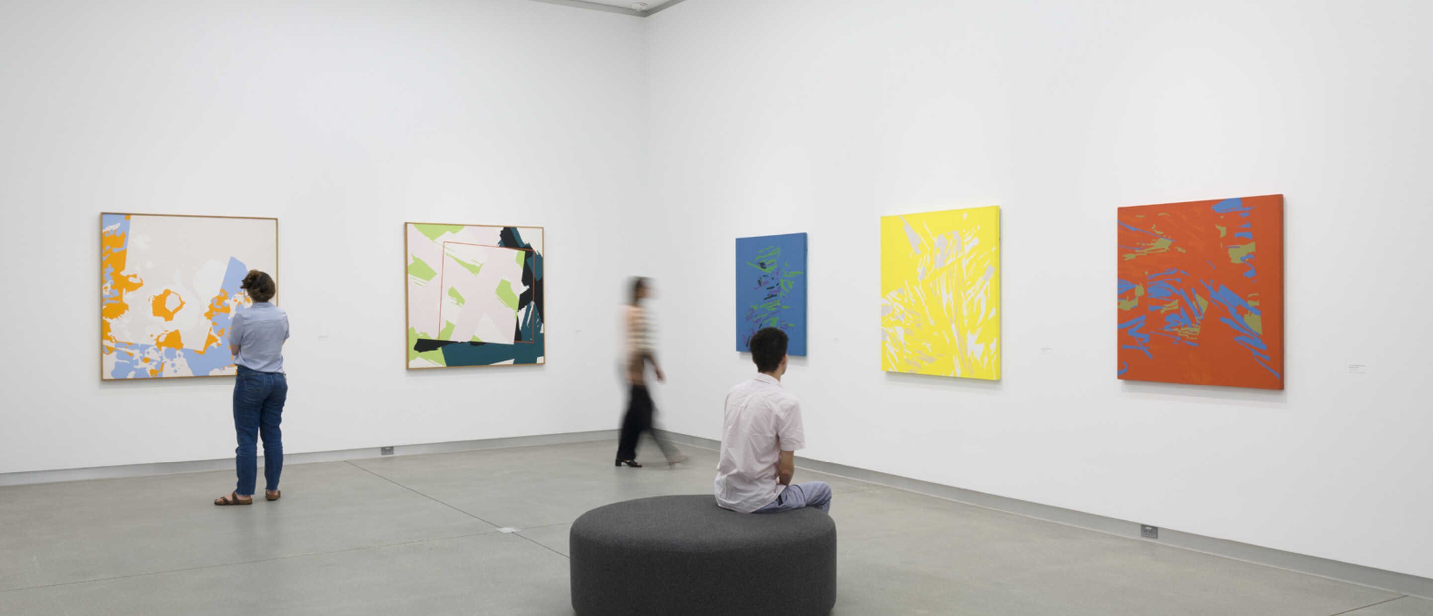 three people looking at colorful paintings on the walls. One person is sitting