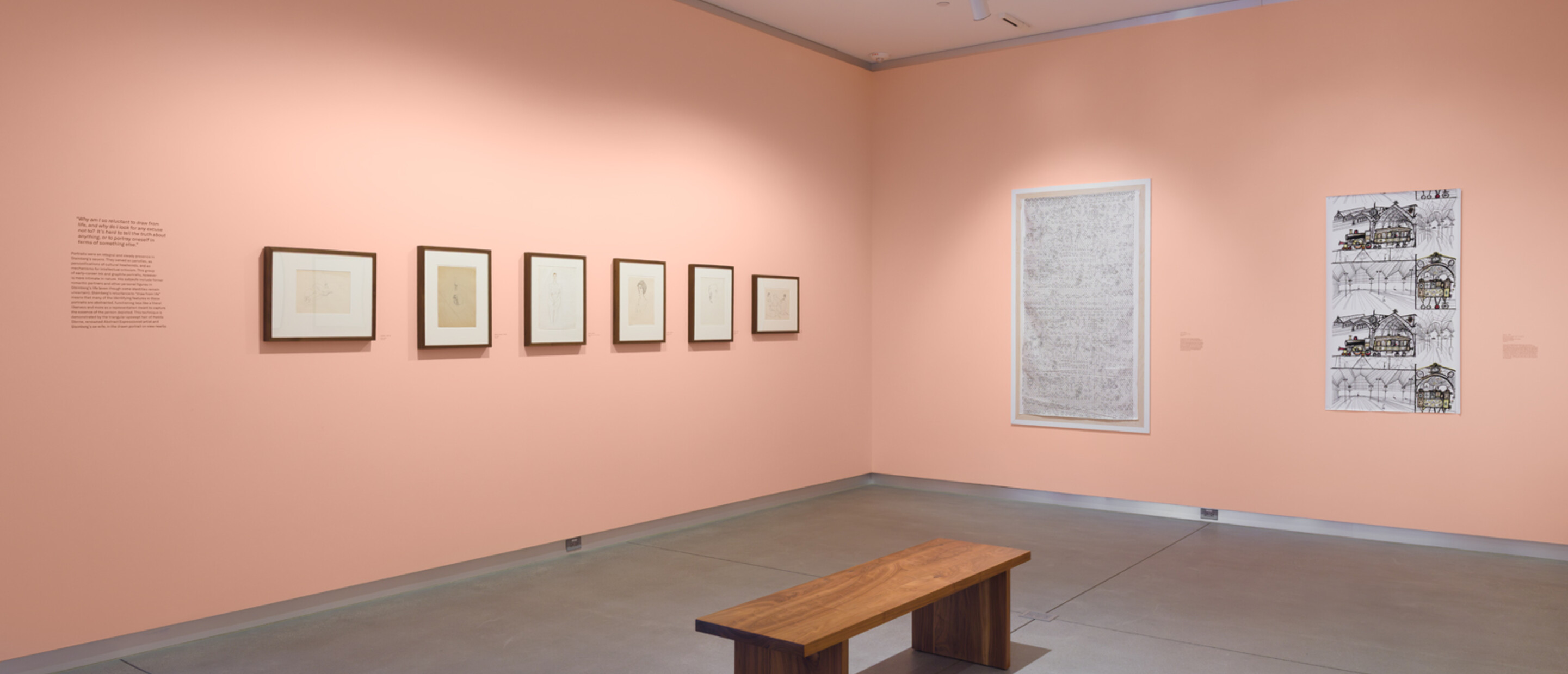 wide shot of a gallery with framed works on pink wall and a wooden bench