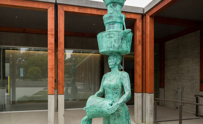 Tall bronze outdoor sculpture of female figure holding stacked pots above while pouring water from a handheld pot
