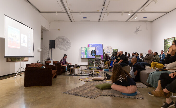 Wideview of an event in a gallery with speaker up front and group of people in the audience