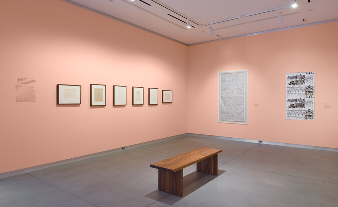 wide shot of a gallery with framed works on pink wall and a wooden bench