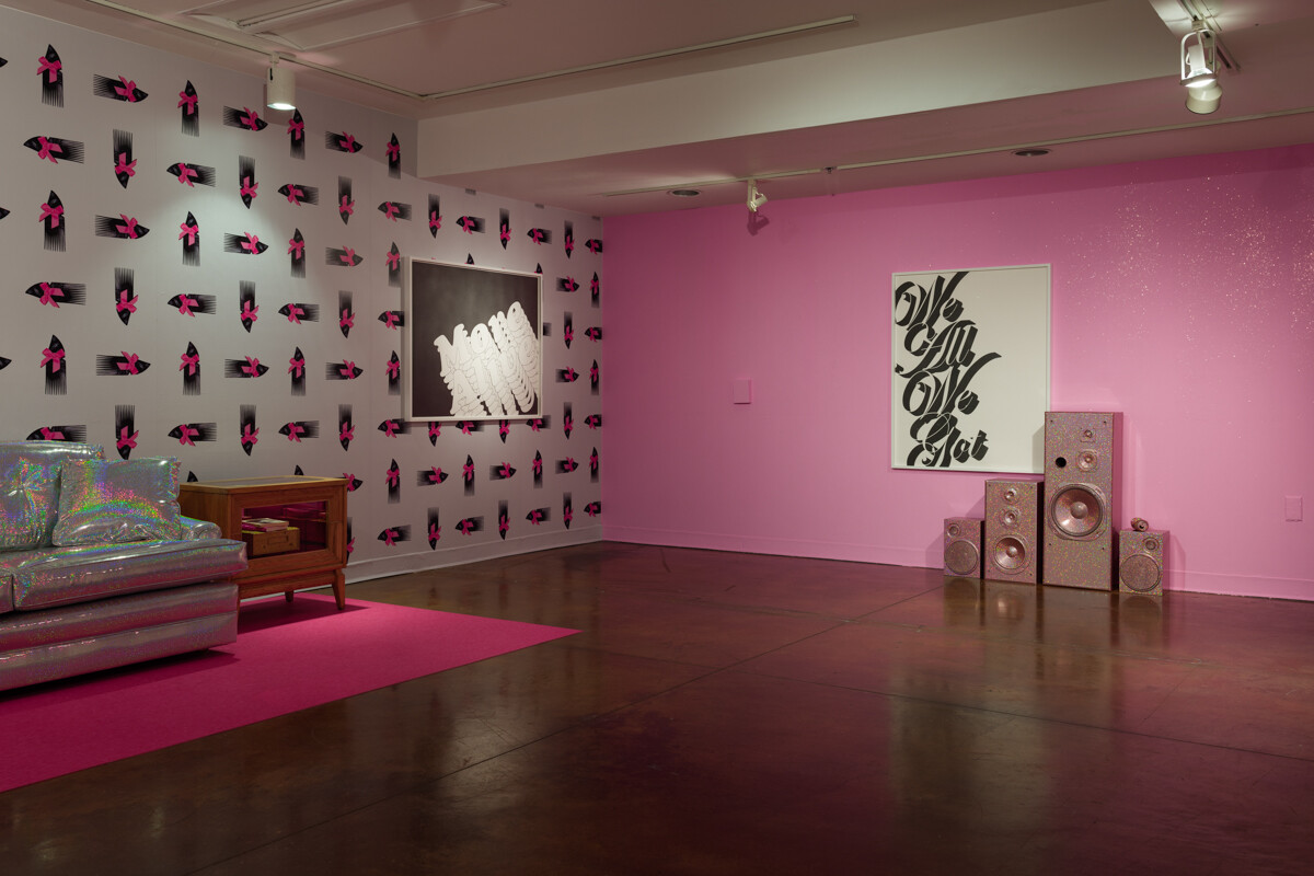 Art installation with 2-dimensional works and a silver couch and pink speakers