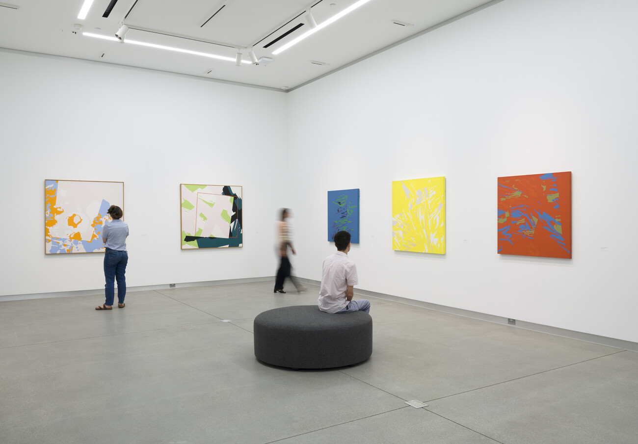 three people looking at colorful paintings on the walls. One person is sitting