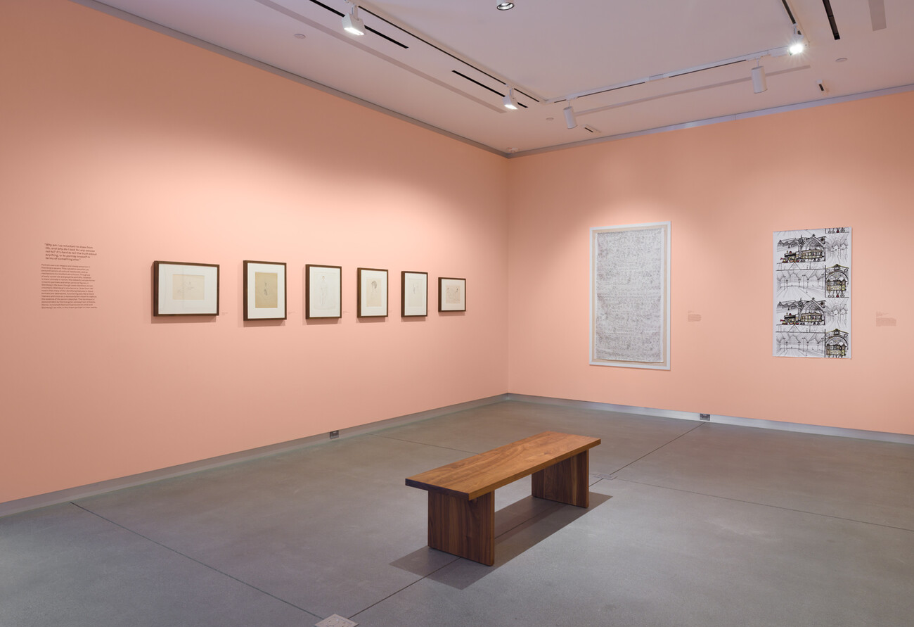 wide shot of a gallery with framed works on pink wall and a wooden bench