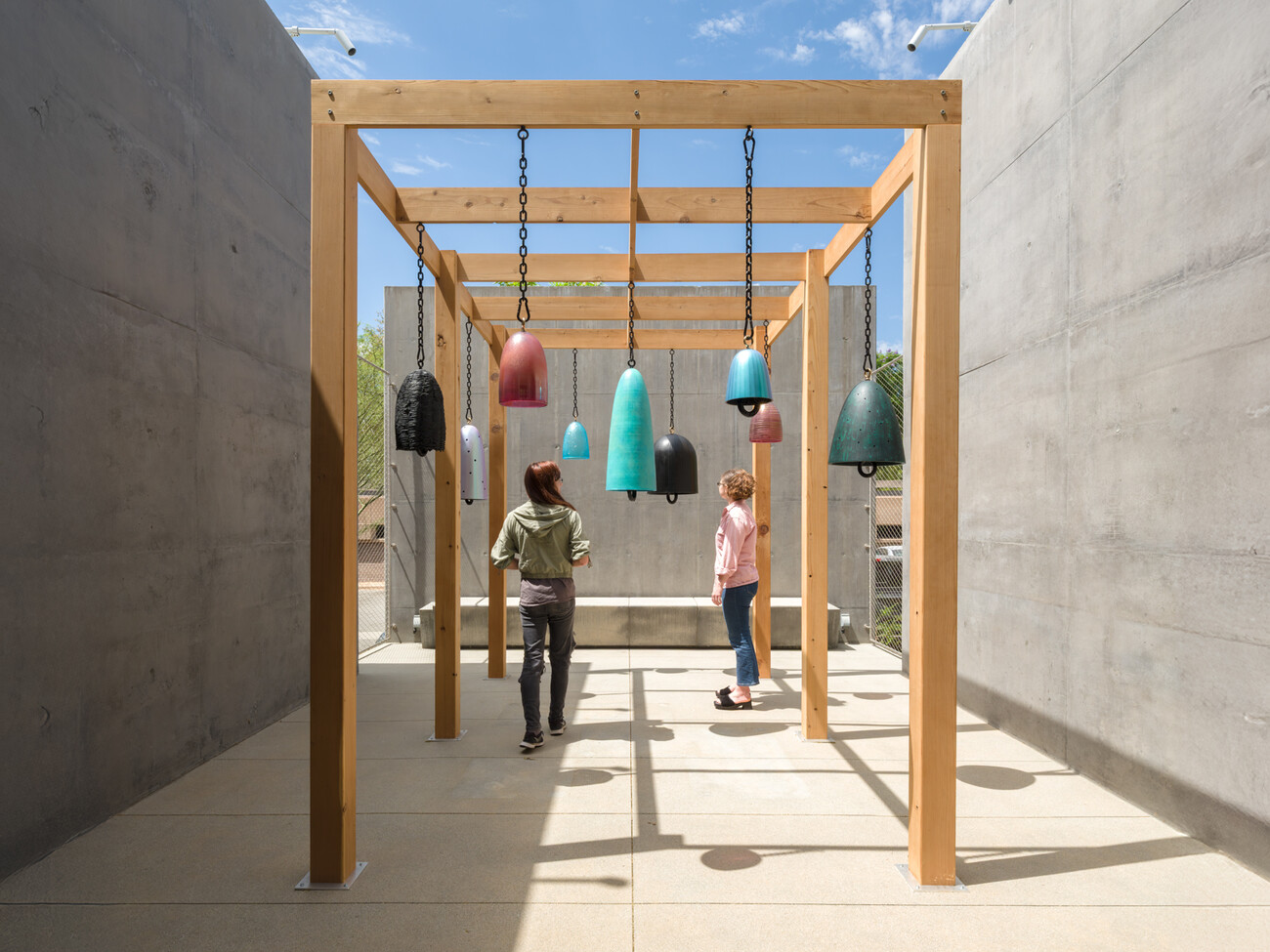 wide shot of two visitors under the array of bronze and glass bells, hung on an outdoor pergola