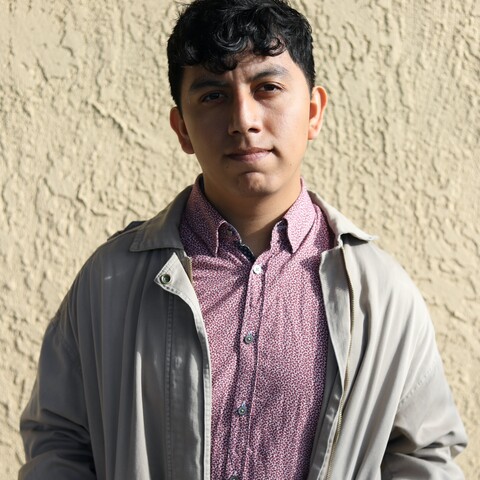 Man wearing purple shirt and beige coat standing in front of stucco wall