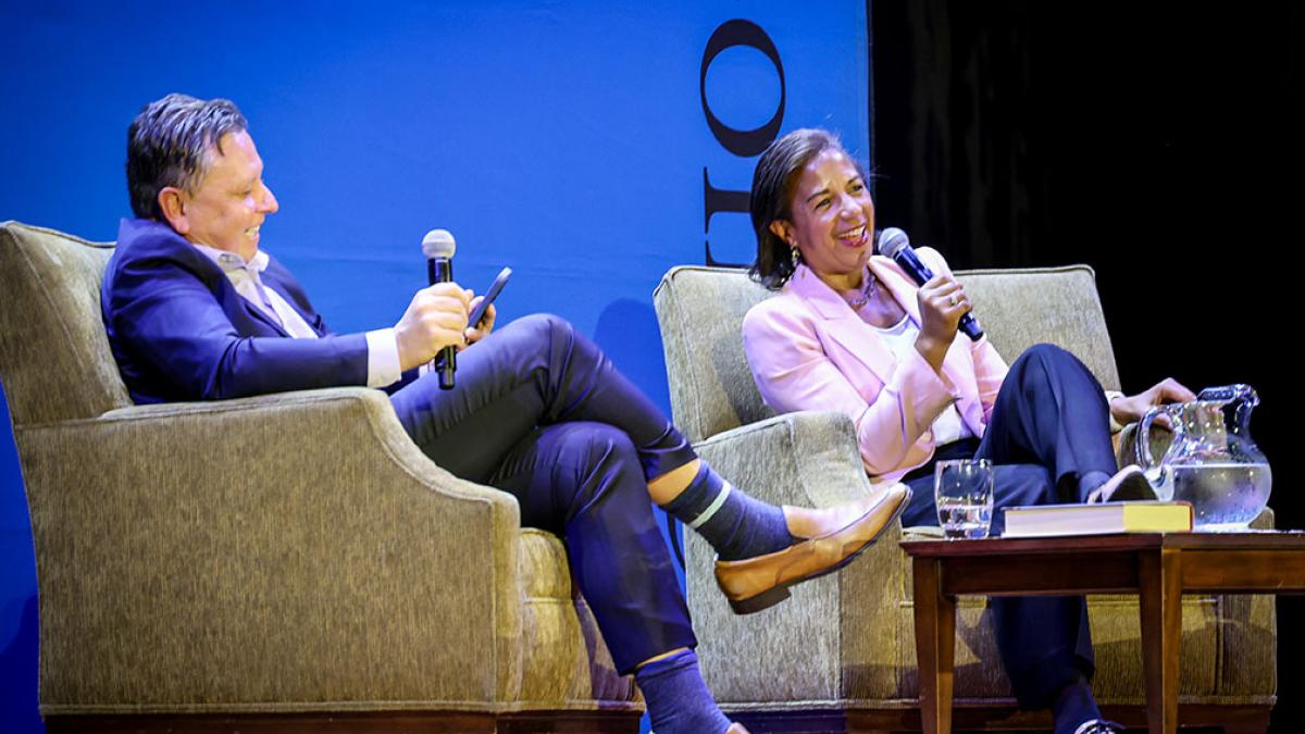 Man wearing blue suit sitting in chair holding microphone and woman wearing pink blazer sitting in chair speaking into microphone