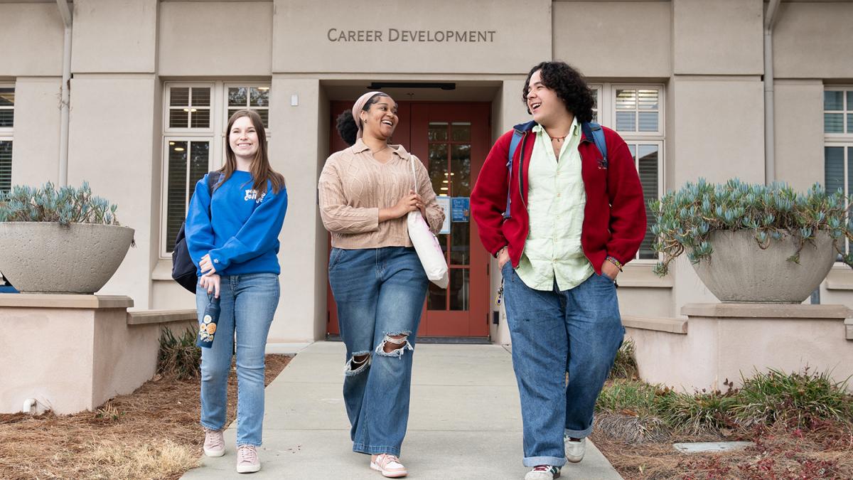 Three students walk on the sidewalk from the Career Development Office front door.