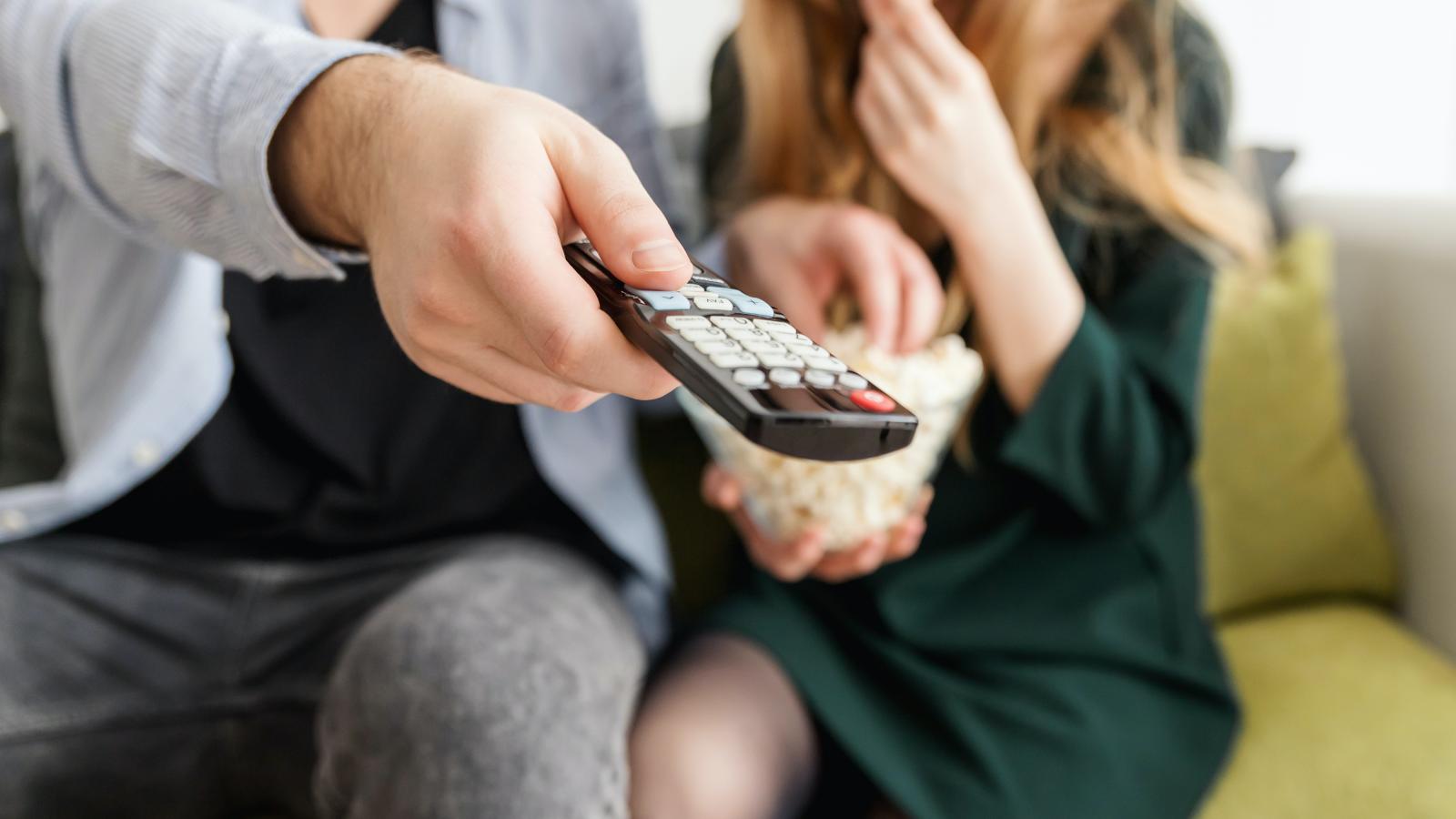 A person using a TV remote and a person eating popcorn on a couch.