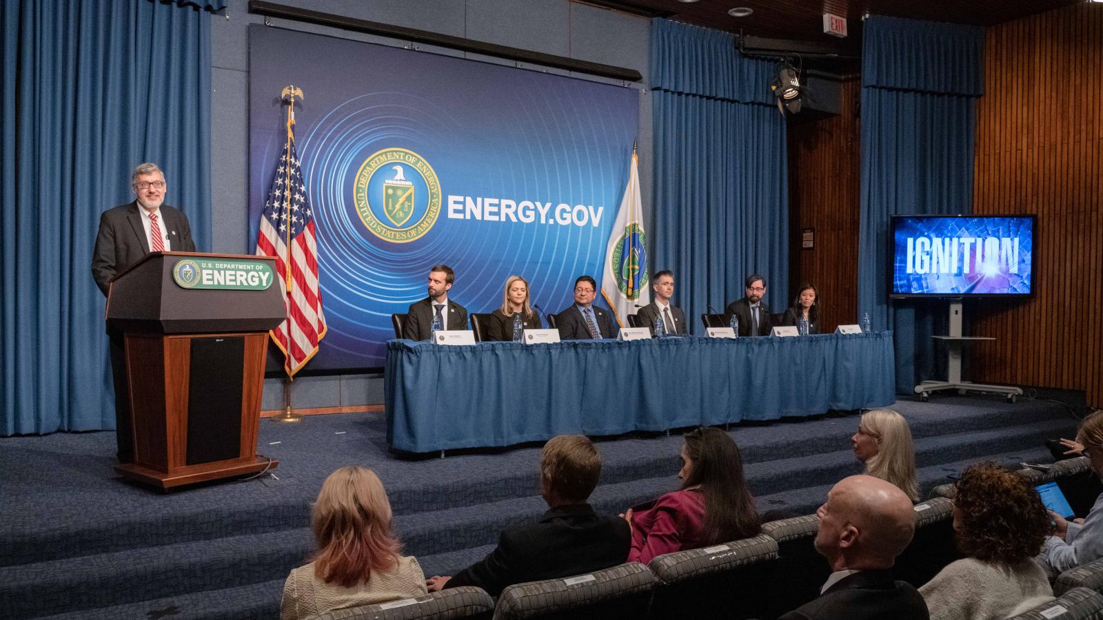 Alex Zylstra '09 and five colleagues are seated at a table; a presenter is standing at a podium during a press conference panel.