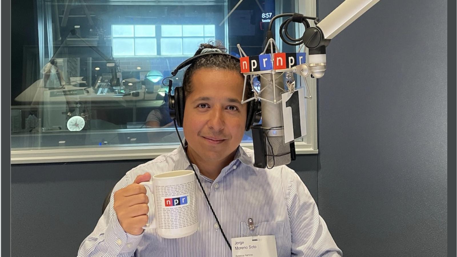 Jorge Moreno poses for photo lifting cup that says NPR behind a microphone