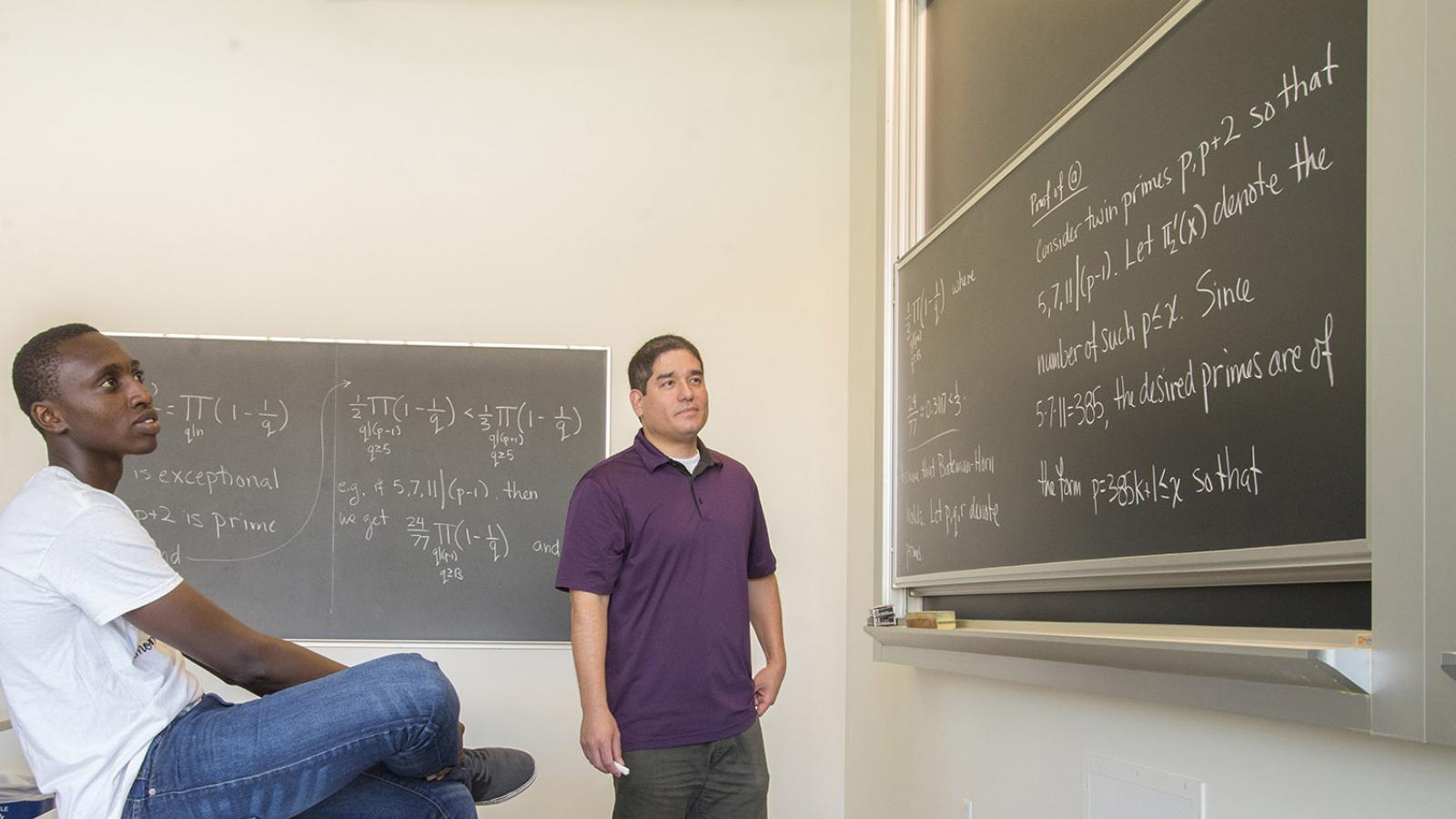 A student is sitting on a table looking at a chalkboard while a professor stands in the middle.