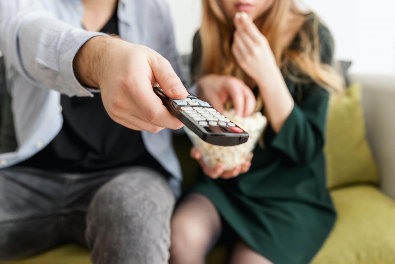 A person using a TV remote and a person eating popcorn on a couch.