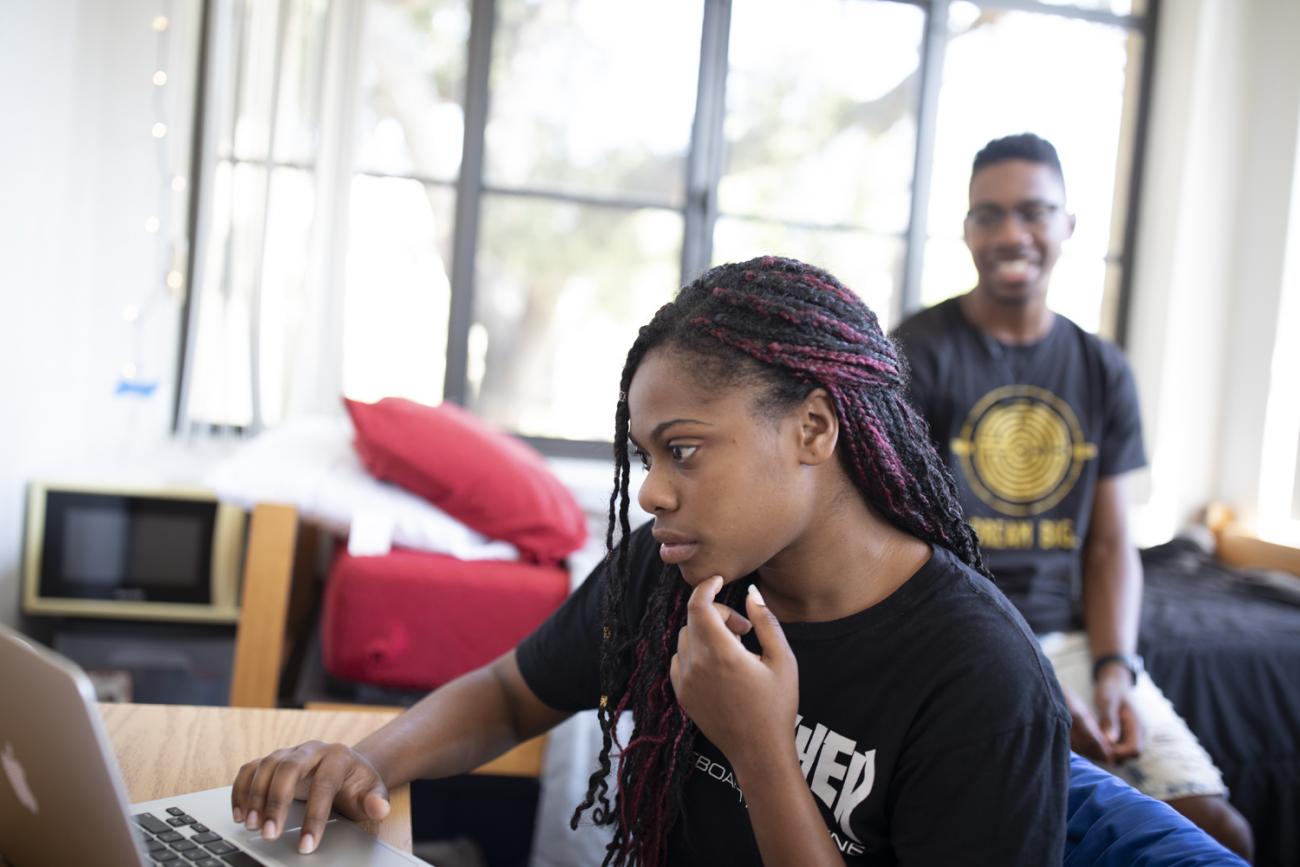 Female student on laptop in dorm room, with male student smiling in the background