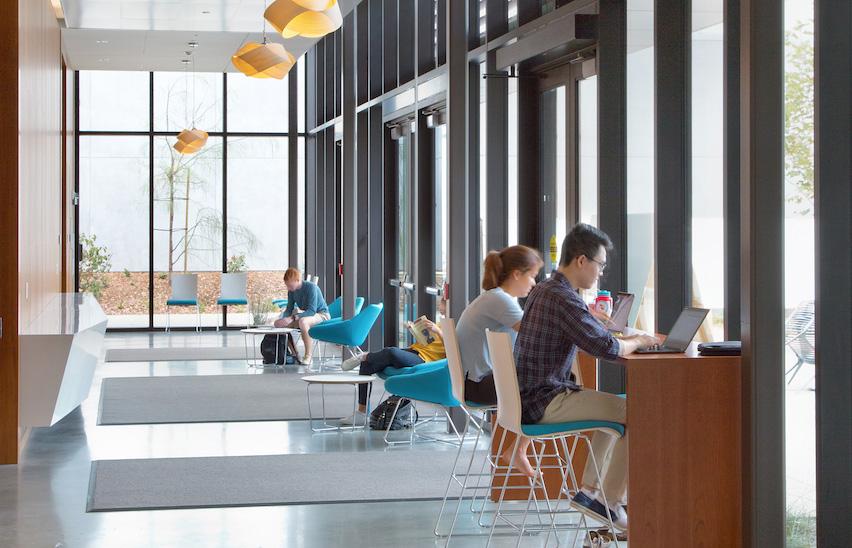 students on laptops on right side of image, hallway with windows