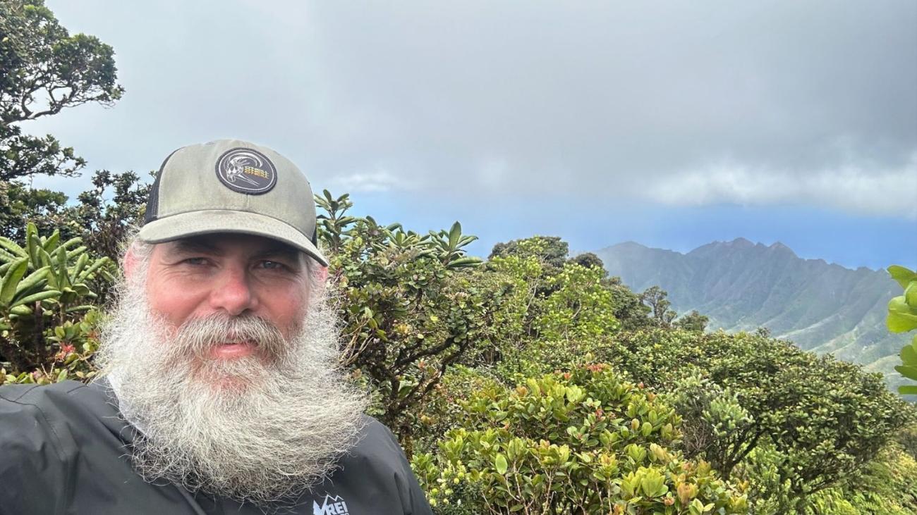 Professor Wallace in a hat with a white beard in front of mountain greenery