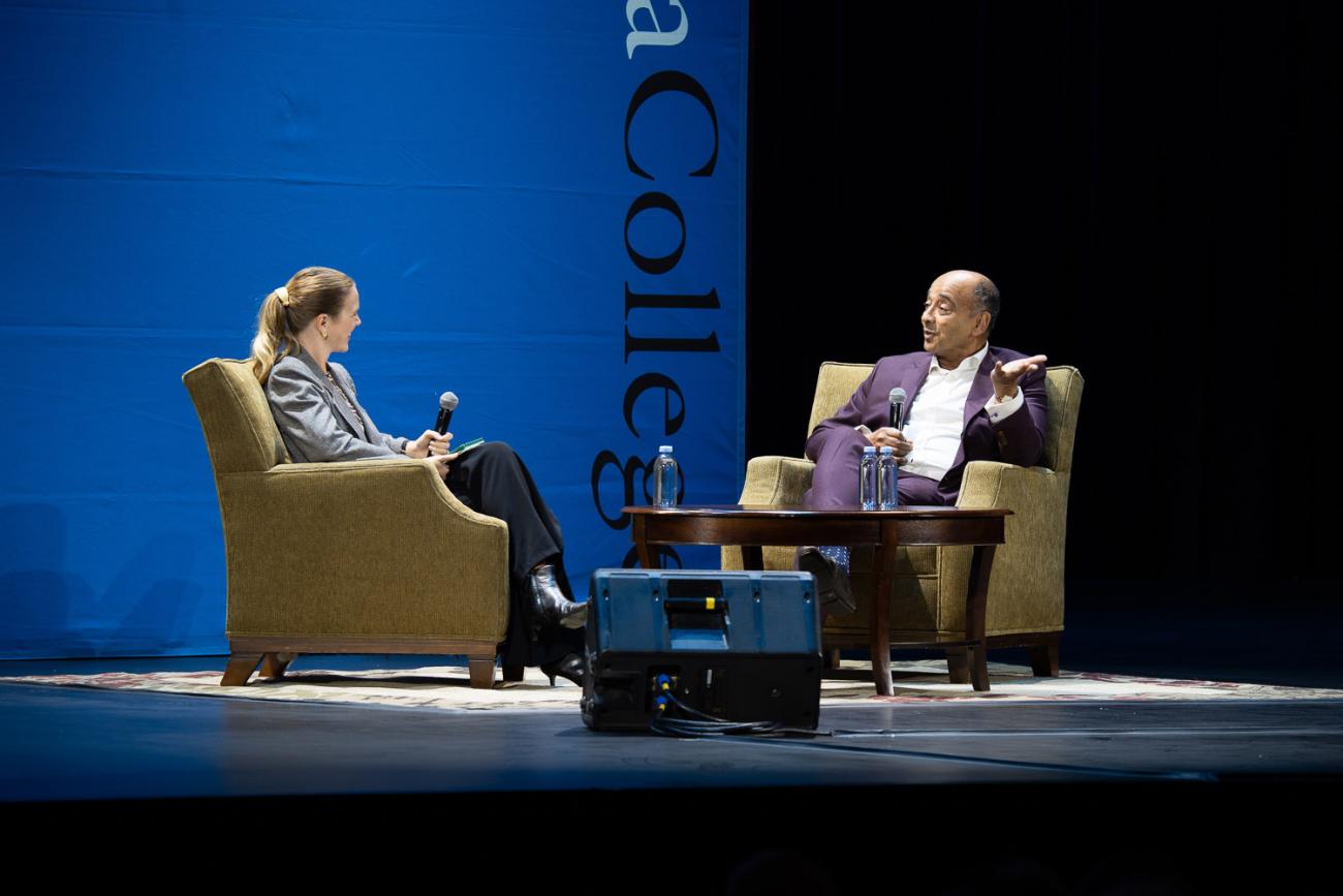 Interviewer and Kwame Anthony Appiah holding microphones, sitting in chairs on a stage with a table between them