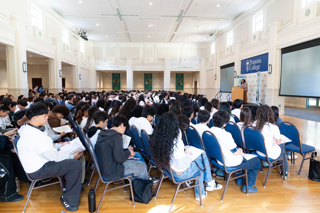 Hundreds of students sit in a ballroom facing a stage with a woman speaking. 