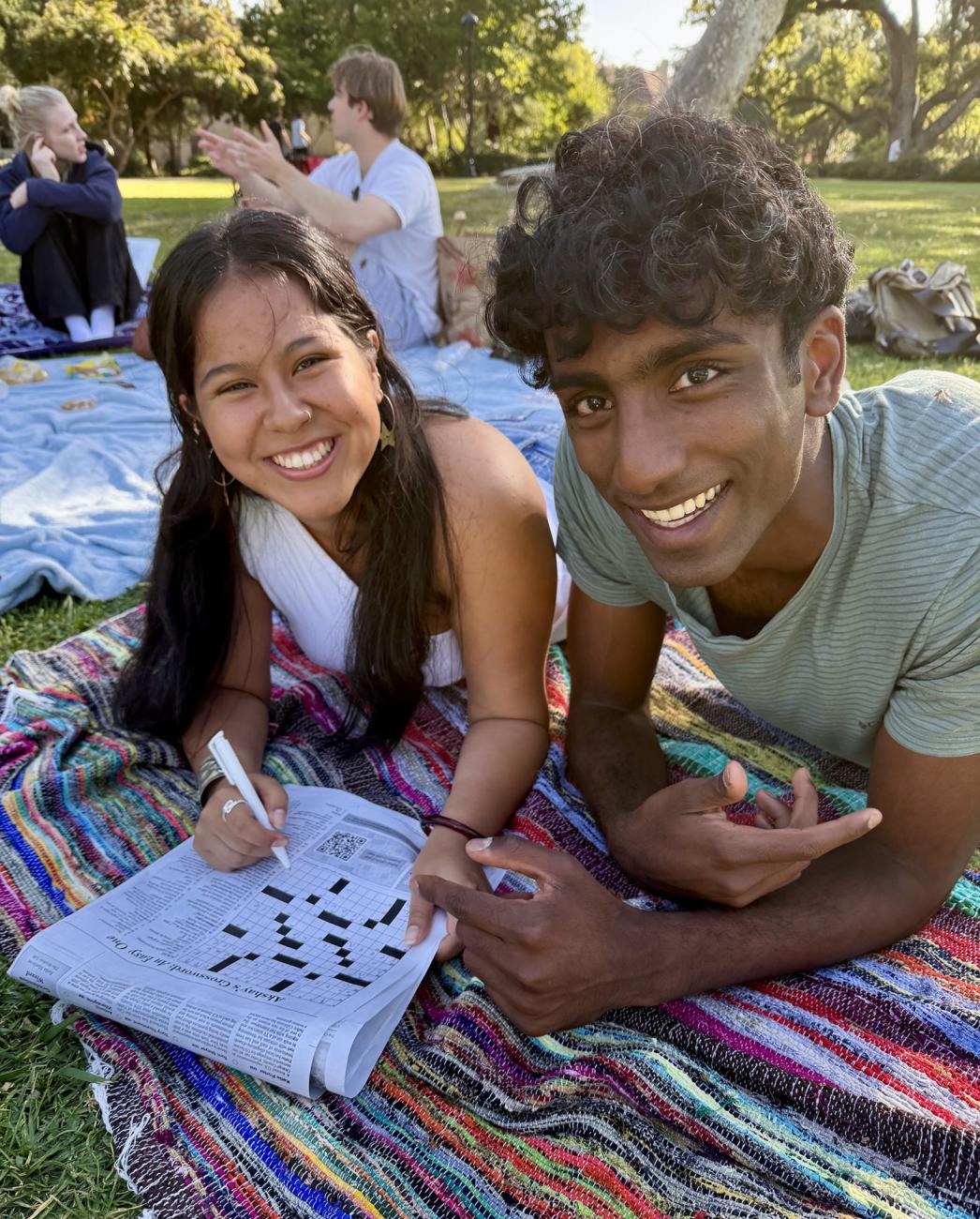 Two students lay on a blanket on Marston Quad working a crossword puzzle.