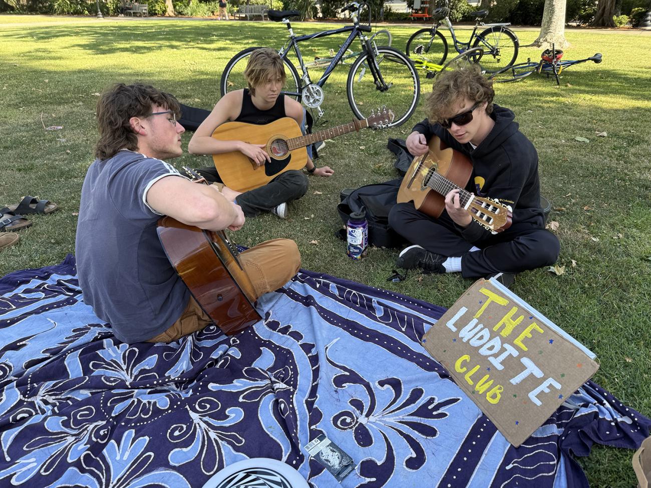 Three students seated on Marston Quad playing guitars.