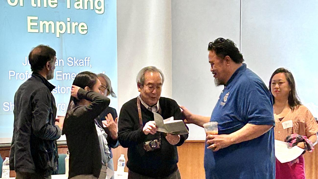 Prof. Samuel Yamashita looks at a book while surrounded by former students and colleagues.