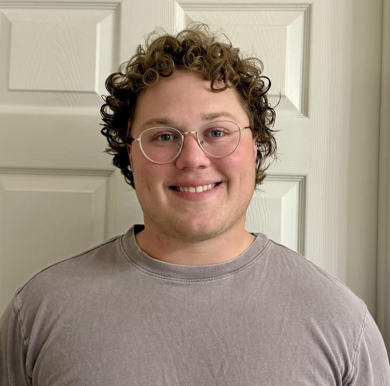  Max Eastman wearing a gray teeshirt stands in front of a white door.