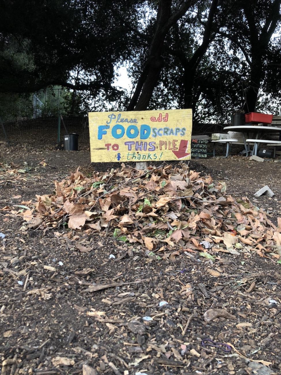Small compost pile with sign that says "Please add food scraps to this pile"