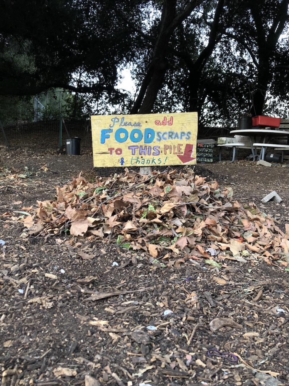 Small compost pile with sign that says "Please add food scraps to this pile"