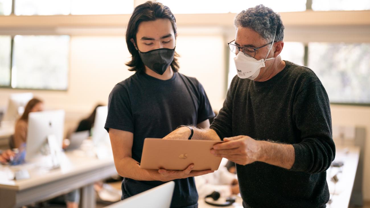 Professor Daniel Martínez talks in class with a student holding a laptop computer.