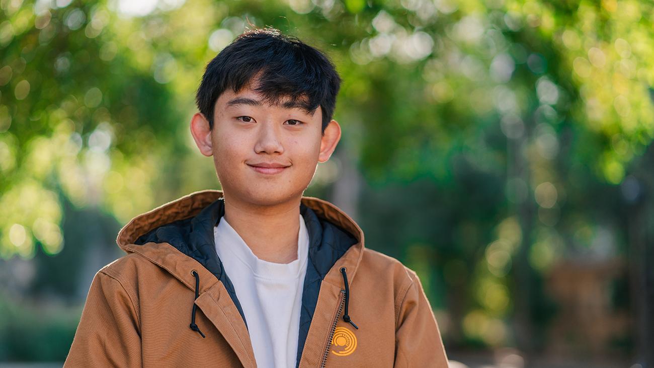 Young man in gray shirt and brown jacket smiling into camera