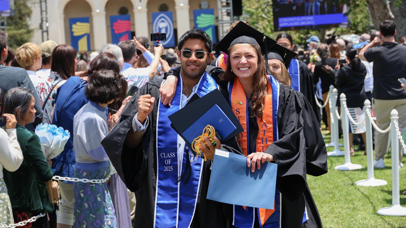 Two graduating Pomona students wearing caps and gowns, walking with arms around each other down the aisle at the completion of Commencement exercises.