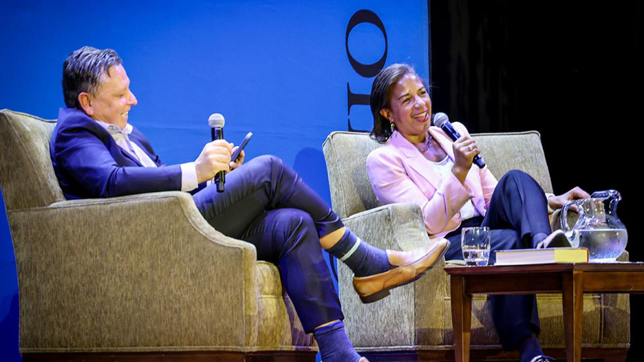 Man wearing blue suit sitting in chair holding microphone and woman wearing pink blazer sitting in chair speaking into microphone