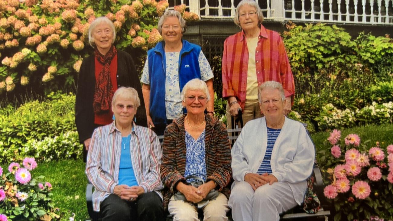 6 women sit and stand in front of a garden filled with flowers