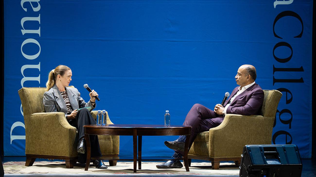 Two adults holding microphones, sitting in chairs on a stage with a table between them