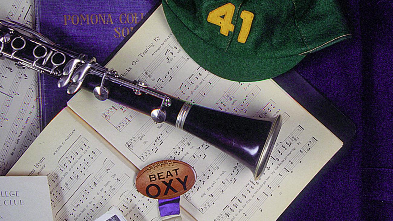 Still life photo of an open music book, a clarinet, a freshman's beanie and other historic paraphernalia related to college songs